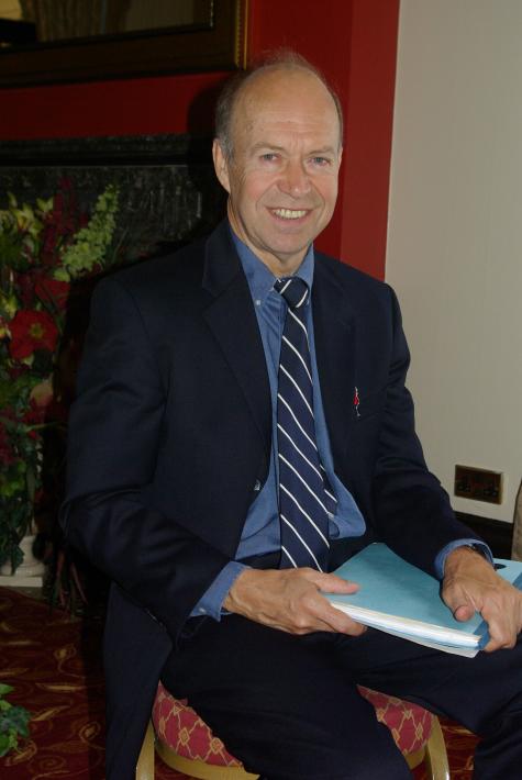 A white older gentleman sits on a chair holding a blue folder wearing a black and blue suit. He smiles warmly at the camera