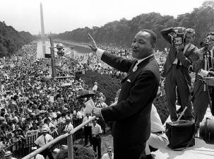 Black and white image of happy African American man waving on a stage with many spectators below and two cameramen filming behind him