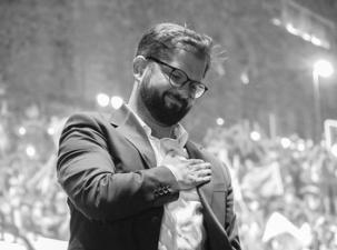 A black and white picture of the president of Chile shows a close up picture of Boric wearing a suit, smiling, and with his hand on his chest.