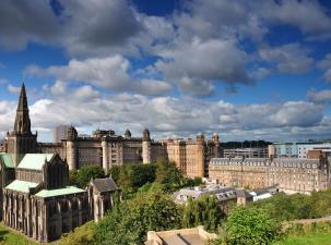 Glasgow city centre cathedral and other old buildings surrounded by trees with a blue sky and white clouds