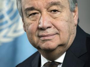 Portrait of an older gentleman with tanned skin, wearing a black suit with a brown tie. He has grey hair and bushy eyebrows, he smiles warmly into the camera