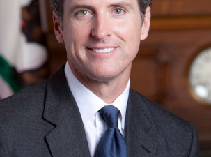Image of California Governor Gavin Newsom, a white man wearing a black suit smils into the camera