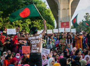 Bangladesh students launch the "Bangla Blockade" demanding the government scrap the discriminatory quotas in public service. A man with no shirt has the demand painted on his back, he is  waving a large Bangladesh flag infront of many other protesters who are all holding signs