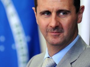 Former Syrian dictator Bashar al-Assad stands in front of a blue background wearing a grey suit, he is looking away from the camera