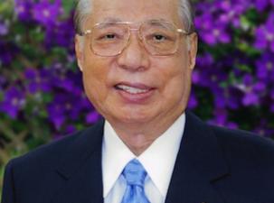 Japanese Buddhist philosopher smiles at the camera wearing a blue suit and tie infront of a purple flowering bush