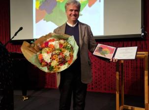 Image of World Beyond War co-founder David Swanson on stage receiving a prize holding a certificate and a large colourful bunch of flowers 