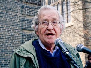 Noam Chomsky is an older white gentleman who is a celebrated linguist, and political analyst. He stands behind two microphones delivering a speech outside.