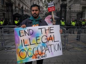 A man stands infront of metal railings at a protest for Palestinian rights. He is holding a sign that says 'End the Illegal Occupation Now!'