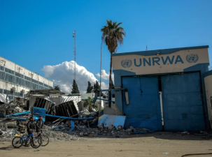 Large blue doors with the letters UNRWA painted above it with the United Nations symbol either side stand surrounded by rubble and destroyed buildings. Two men pushing bicycles are walking by.