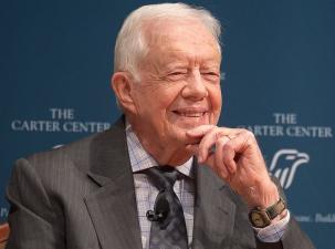 Photo of USA ex-president Jimmy Carter in his 90s. He is wearing a grey suit and smiling in front of a large sign for the Carter Center 