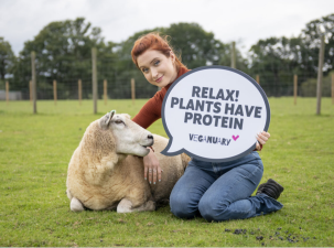 Photoshoot to promote the Veganuary initiative. A lady with orange hair leans on the floor next to a sheep in a large open field. She holds a sign that says 'Relax! Plants have protein.'
