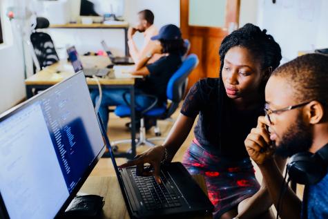 AnArfican-American man and woman sit at a computer and laptop while the women is helping to show him something. There are other desks behing them in a library type setting.