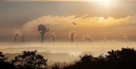 Many dark plumes of smoke can be seen above factories and powerplants. Electric towers and cables can be seen in the background. The air is covered in an orange smog.