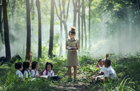 A slim Asian women wearing beige outdoor clothes stands holding a book infront of 6 children who are st with their legs crossed on the floor, two of the children has their arms raised as if to answer a question. They are in a beautiful forest setting surrounded by trees and plants with the sun shining through.
