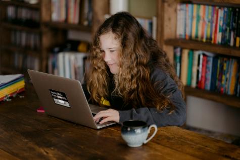 A young girl sits in a library at a desk looking excitedly at her laptop screen