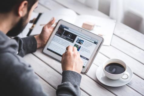 Man with dark hair sitting at a table concentrating on his tablet drinking a coffee