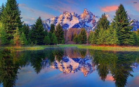 A beautiful snowy mountain range sits behind rows of pine trees. With the orange of the sunset captured in the white clouds infront of a blue sky. The whole image is perfectly reflected into a bright blue still lake.