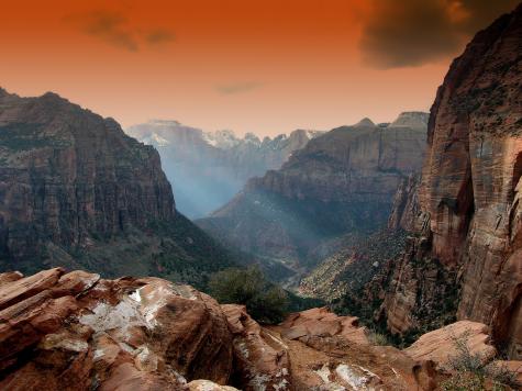 Four large steep rocky mountains are viewed from an over hang. The sky in the background is orange and shines through onto the mountains.