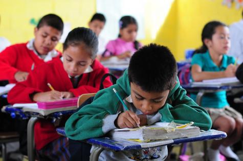Young school children, boys and girls, sit at their desks in Villa Nueva, Guatemala working hard. The children all he dark hair and are not wearing a uniform. They work on old desks with tattered books
