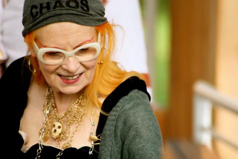 Cultural icon Vivienne Westwood sports her trademark orange hair at an Active Resistance to Propaganda event. She is smiling, looking away from the camera, wearing an assortment of necklaces and a grey headband that reads 'chaos'