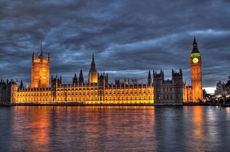 The Bristish House of Parliament and Big Ben under a cloudy night sky. The building and clock tower is lit up in an orange glow. The river Thames flows infront with the light reflecting on it.