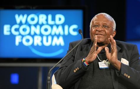 An older gentleman of colour wears a black suit and holds his hands almost together infront of his chest whilst he is talking