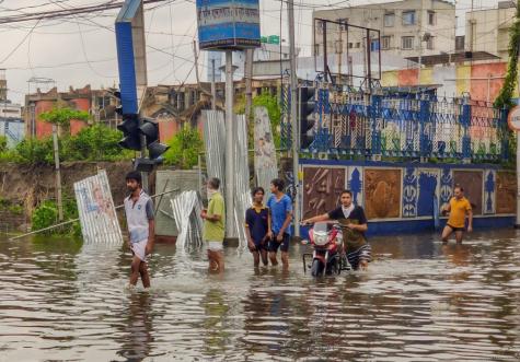 Six men and boys walk through dark knee high water on a road. One man is pushing a motorbike. In the background there are tall dirty looking buidings, there is a broken wall, and traffic lights and cables have dangerously fallen over.