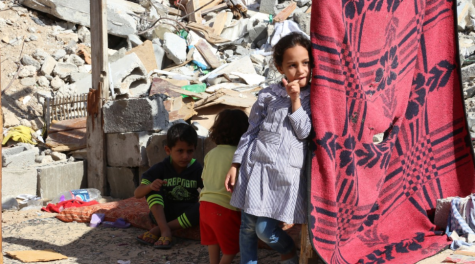 A girl and her two younger siblings stand in the rubble of a destroyed building in Gaza. There is a red sheet hanging to provide them with shade