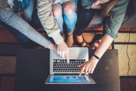 Image from above of three women sitting on a sofa around a silver laptop. You can not see the girls lfaces, only their legs and their arms which are pointing at the screen