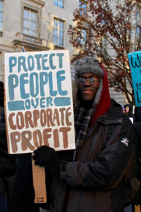 A young black man wearing a red hat and black coat is at a march in Washington. He holds a white banner which reads ' Protect people over corporate profit'