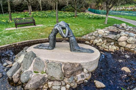 Metal sculpture by Josefina de Vasconcellos. A man and woman on their knees embrace each other. The sculpture sits ontop of a large stone and concrete circle base in the centre of a pond