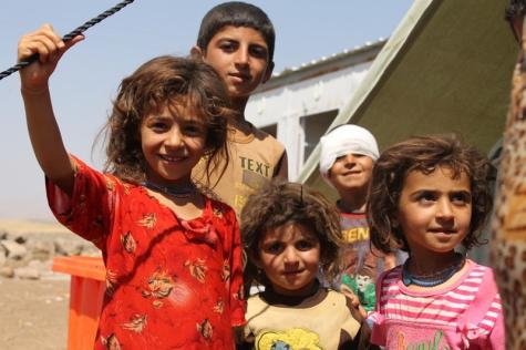 5 young Iraqi children, boys and girls stand smiling looking into the camera. One has a bandage on his head. The children are refugees living at a camp in Syria