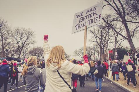 A large group of people protest in Washington, DC with their backs to the camera. One woman hold a large white sign that reads 'We are better than this.'