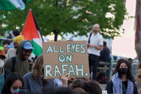 People gather at a pro-Palestine protest. There are Palestinian flags in the background and someone holds a cardboard sign which reads 'All eyes on Rafah'