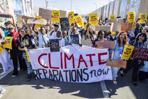 Climate reparations protest at COP27 in Egypt. A group of people holding yellow signs saying 'show us the money' stand behind a large white banner which reads 'Climate reparations now'