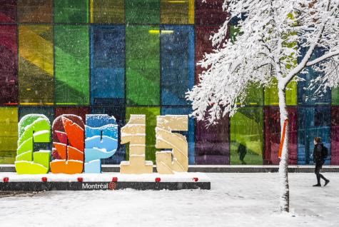 The colourful COP15 sign sits infront of a wall filled with coloured glass, snow is falling and it covers the ground and a large tree in the foreground