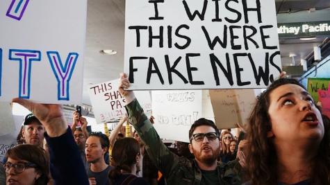 Image of protesters holding signs. The main sign is being held up by a man wearing glasses, it is large and white and reads 'I wish this were fake news'