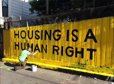 Housing Is A Human Right grafitti - A man paints large black graffiti on a yellow wooden fence