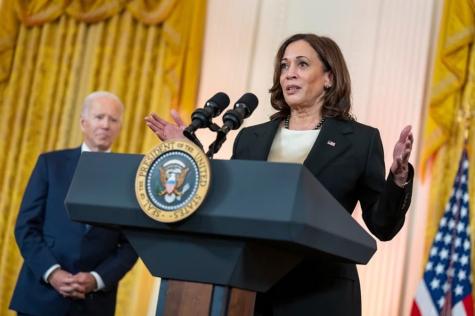 Kamala Harris stands behind a podium delivering a speech infront of a yellow background with a U.S. flag. Biden stands off to the side with his hands clasped