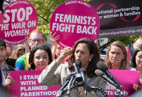 Wearing a grey suit Kamala Harris delivers a speech with her fist in the air at a women's reproductive rights rally. There is a crowd of women behind her holding large pink signs that say 'feminists fight back' and 'stop the war on women'