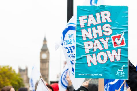 Image of NHS protesters fighting for fairer pay at a demonstration in London. In the foreground is a blue poster which says 'NHS fair pay now' and Big Ben is in the background