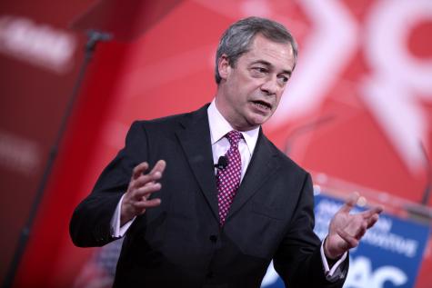 Nigel Farage speaking at the 2015 Conservative Political Action Conference. He stands infront of a red background wearing a black suit holding his hands open and looking away from the camera