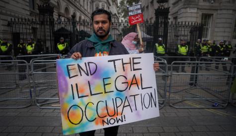 A man stands infront of metal railings at a protest for Palestinian rights. He is holding a sign that says 'End the Illegal Occupation Now!'