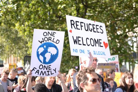 Protesters gather outside to support the arrival of refugees. There are two larg white signs one reads 'One world, one family' and the other says 'Refugees welcome noone is illegal.'