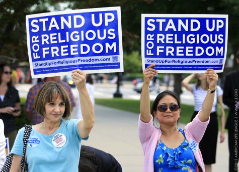 Two women wearing blue are at a rally supporting religious freedom, they both hold up larg blue posters that say 'Stand up for religious freedom'