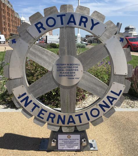 A Rotary Coin Collector in the UK sits infront of a field. There is a large silver cog and below it a slot for donators to leave money. The words Rotary International are written in large letters around the cog.