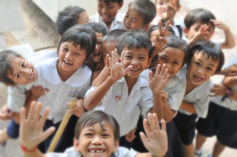 A group of school children wearing white uniforms gather around the camera smiling, giving peace signs and holding their hands in the air