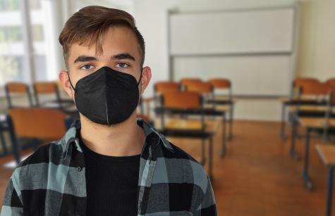 A student wearing a grey shirt stands in a classroom wearing a black Covid mask