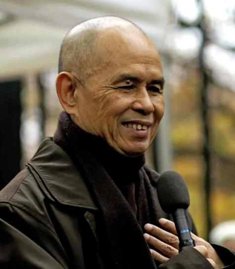 Buddhist monk Thich Nhat Hanh wears a black sweatshirt and jacket, he is looking down into a microphone smiling