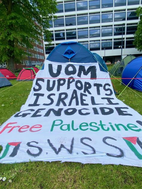 A large white cloth painted with Palestinian solidarity messages is spread out over a tent in the University of Manchester protest encampment. 